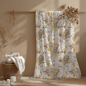 May include: A white and beige patterned curtain with watercolour illustrations of owls on branches with autumn leaves. The curtain hangs on a wooden rod. A woven basket and candles are in the foreground.