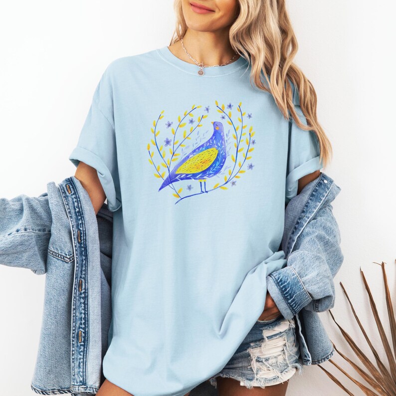 Puede incluir: Camiseta azul claro con una ilustraci&oacute;n estilizada de un p&aacute;jaro. El p&aacute;jaro es azul y amarillo, rodeado por una corona de hojas amarillas y flores moradas. La camiseta tiene un corte holgado y casual.