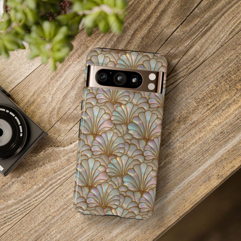 May include: A smartphone case with a pearlescent, fan-shaped pattern in gold and iridescent colors. The case is on a wooden surface, with a camera and plant in the background. The case is designed for a smartphone.