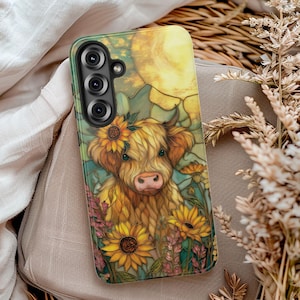 May include: A phone case featuring a Highland cow illustration with sunflowers and a large sun in the background. The case has a green border and a camera cutout. The cow has a flower crown and is surrounded by colorful flowers.