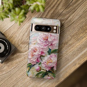 K&ouml;nnte beinhalten: Eine wei&szlig;e Handyh&uuml;lle mit einem Blumendesign mit rosa Pfingstrosen und gr&uuml;nen Bl&auml;ttern. Die H&uuml;lle hat eine goldene Kameraerh&ouml;hung mit drei Objektiven.