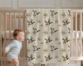 May include: A beige baby blanket with a repeating pattern of flying ducks, reeds, and the name "Reed". The ducks are depicted with brown, green, and blue feathers. The blanket is draped over a wooden crib.