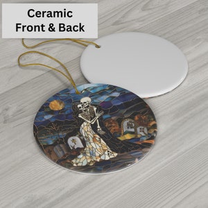 May include: Ceramic ornament featuring a stained glass style image of a skeleton couple embracing in a graveyard setting. The ornament is round and has a white back.