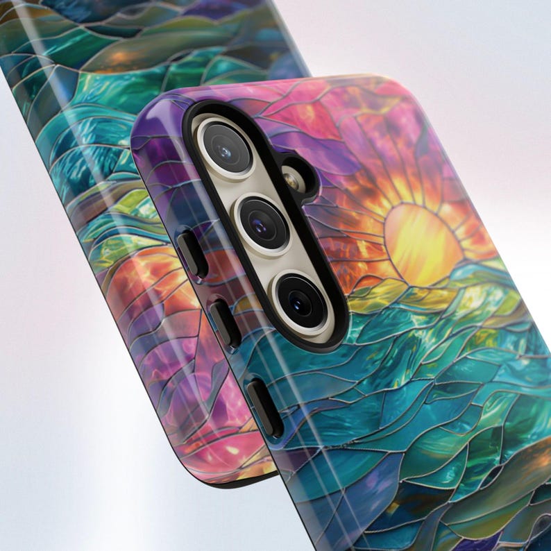 Pu&ograve; includere: Una custodia per telefono colorata con un designo a vetri colorati che rappresenta un tramonto su un corpo d'acqua. La custodia ha un ritaglio per la fotocamera nero con tre obiettivi.