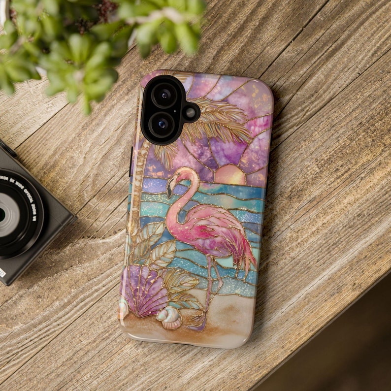 Puede incluir: Funda de tel&eacute;fono con un flamenco rosa en una playa con puesta de sol. El dise&ntilde;o de mosaico presenta palmeras, conchas marinas y una paleta de colores azul y morado. La funda est&aacute; sobre una superficie de madera.