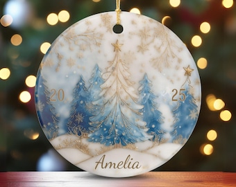 Puede incluir: Adorno navideño redondo con una escena invernal. Presenta árboles azules, copos de nieve y el nombre "Amelia". También están presentes los números "20" y "25". Se adjunta un hilo dorado para colgarlo.