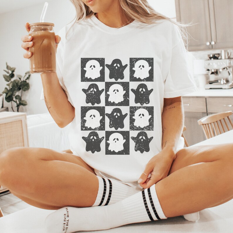 May include: White t-shirt with a black and white checkered pattern of ghosts. The ghosts are in a distressed, vintage style.