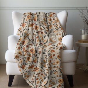 May include: A beige throw blanket with a pattern of owls and autumn leaves in shades of brown, orange, and yellow. The blanket is draped over a white armchair.
