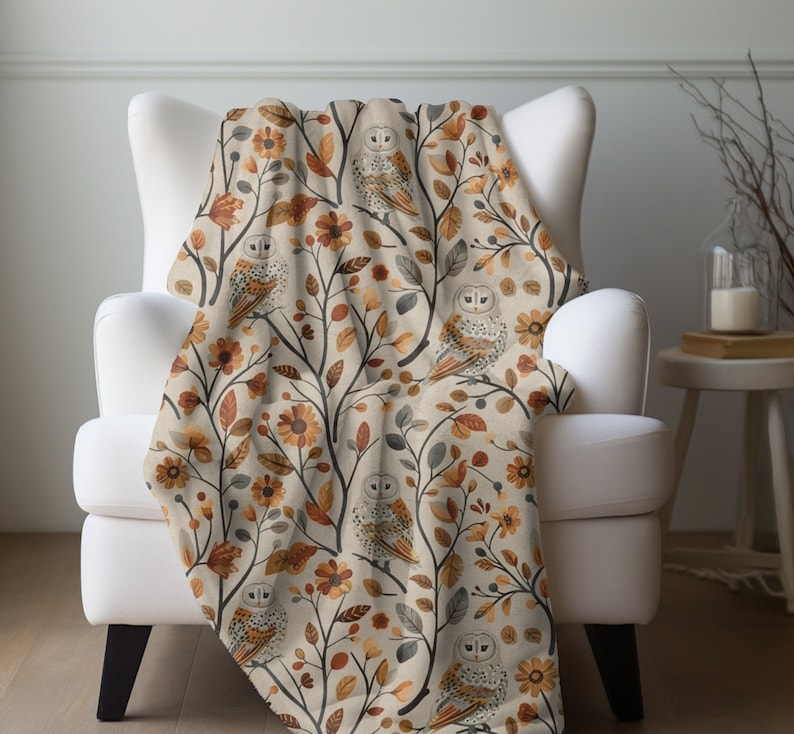May include: A beige throw blanket with a pattern of owls and autumn leaves in shades of brown, orange, and yellow. The blanket is draped over a white armchair.