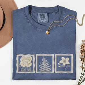 May include: A blue t-shirt with a floral stamp design. The shirt features three vintage-style stamps with a rose, fern, and flower. A gold star pendant necklace rests on the shirt. The tag reads "COMFORT COLORS" and "XL - TG - EG".