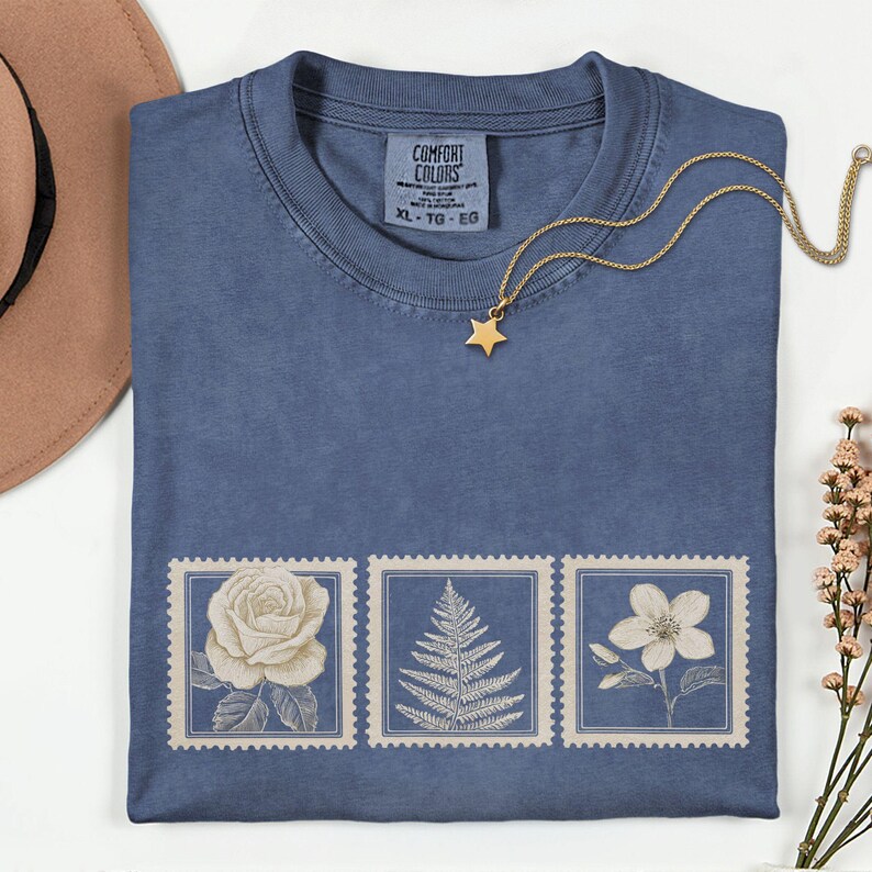May include: A blue t-shirt with a floral stamp design. The shirt features three vintage-style stamps with a rose, fern, and flower. A gold star pendant necklace rests on the shirt. The tag reads "COMFORT COLORS" and "XL - TG - EG".