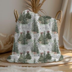 May include: A white blanket with a repeating pattern of watercolor Christmas trees, each adorned with a red bow and a gift. The words "Merry Christmas" are scattered across the design, along with snowflakes. The blanket is folded in a wicker basket.