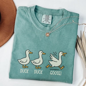 May include: A teal Comfort Colors t-shirt featuring a graphic of three ducks, with the text "DUCK DUCK GOOSE!". The first two ducks are standing, while the third is running. A silver necklace with a pendant is draped across the shirt.