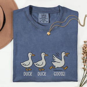 May include: A blue Comfort Colors t-shirt with a playful "Duck Duck Goose!" graphic. The design features three cartoon ducks, with the third one running. A gold star pendant necklace and a brown hat are also visible.