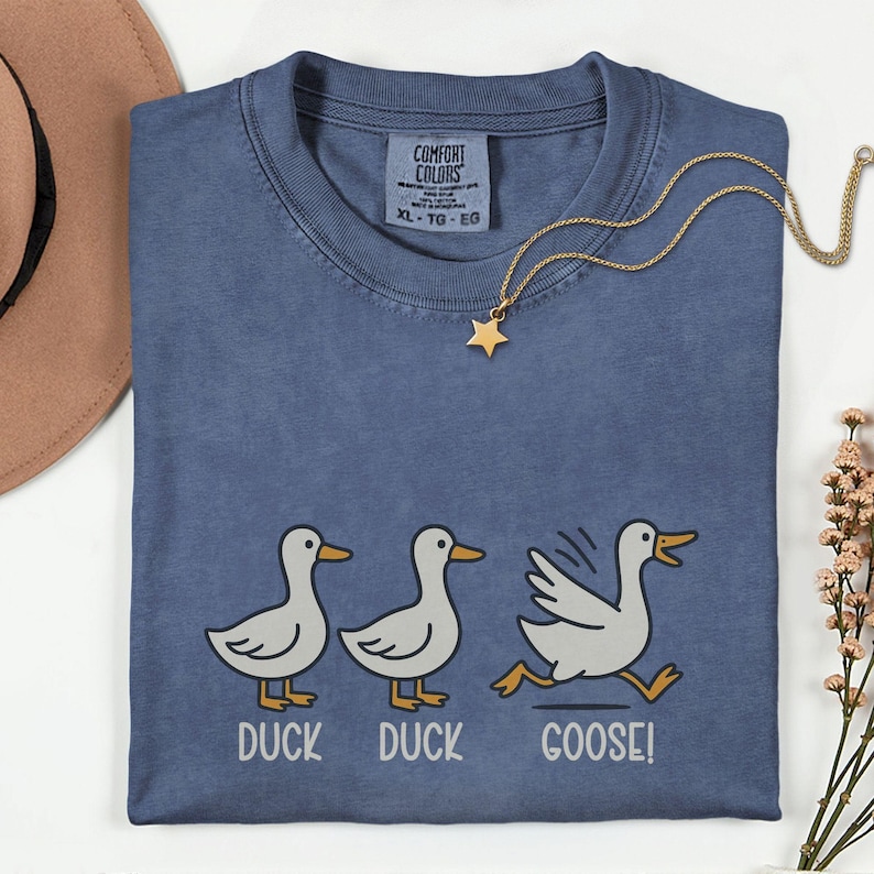 May include: A blue Comfort Colors t-shirt with a playful "Duck Duck Goose!" graphic. The design features three cartoon ducks, with the third one running. A gold star pendant necklace and a brown hat are also visible.