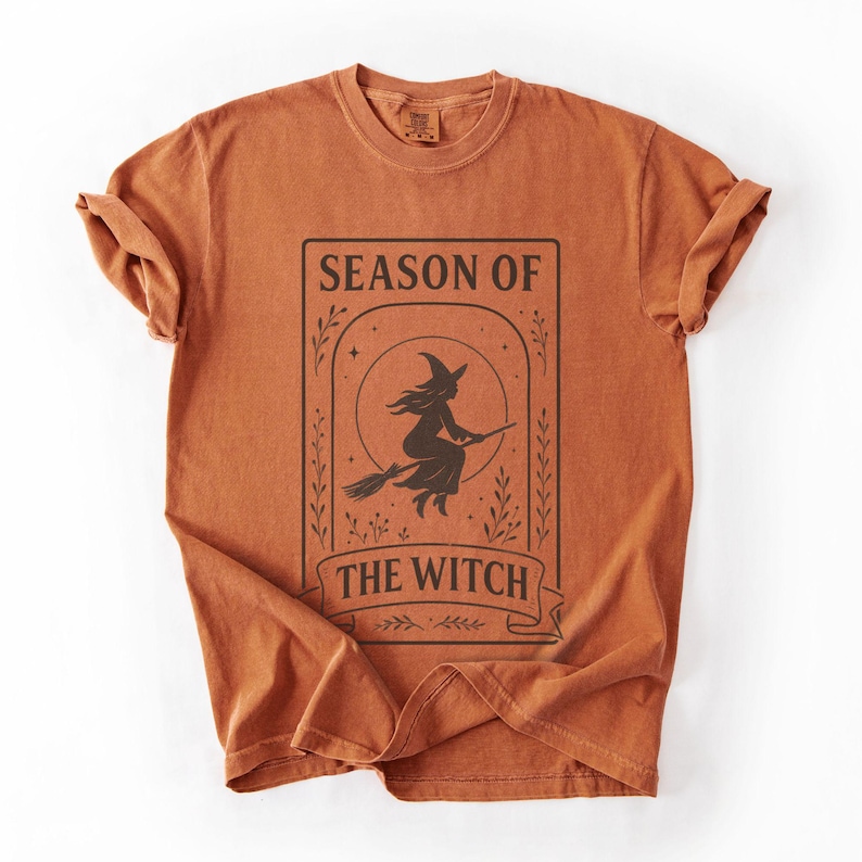 May include: Burnt orange t-shirt with a black graphic of a witch on a broomstick inside a rectangular frame. The text "SEASON OF THE WITCH" is above and below the witch illustration. The shirt has rolled-up sleeves.