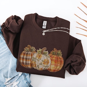 May include: A dark brown crewneck sweatshirt featuring a graphic of three pumpkins. The left pumpkin has a grey and orange plaid pattern, the center pumpkin has a yellow floral print, and the right pumpkin has an orange and yellow plaid pattern.