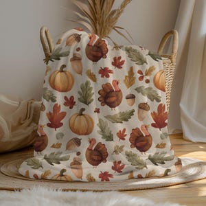 May include: A beige blanket with a Thanksgiving theme, featuring illustrations of pumpkins, turkeys, acorns, and fall leaves in shades of orange, brown, and green. The blanket is folded in a wicker basket.