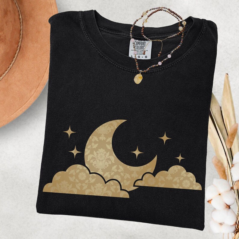 May include: Black t-shirt with a gold crescent moon and cloud design. The moon features a damask pattern, and stars are scattered around the moon. A necklace with a pendant rests on the shirt. The tag reads "Comfort Colors".