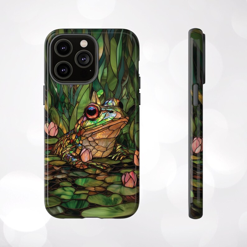 May include: A green, brown, and blue stained glass phone case with a frog sitting on lily pads. The phone case has a cutout for the camera lens.