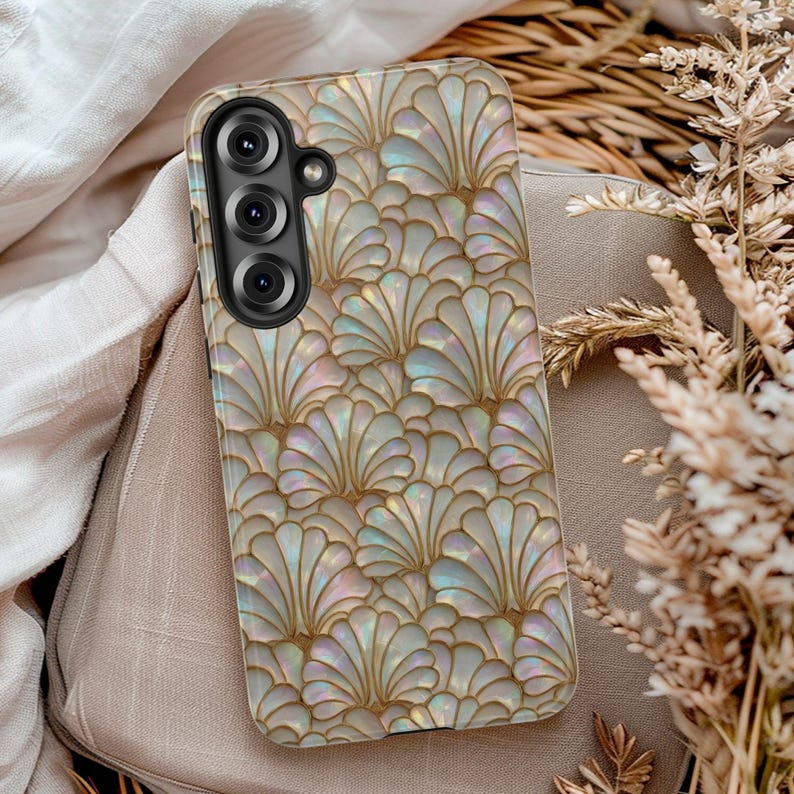 May include: A pearlescent phone case with a repeating fan-shaped pattern in gold and iridescent colors. The case has a camera module with three lenses. The case is resting on a beige surface.