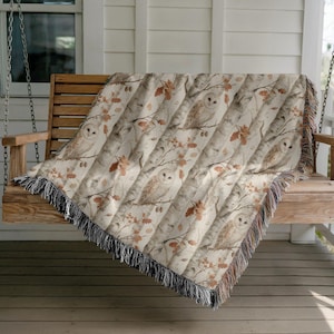 May include: A woven throw blanket featuring a repeating pattern of owls perched on birch tree branches with scattered brown oak leaves. The blanket has a cream-colored background and is finished with decorative fringe along the edges.