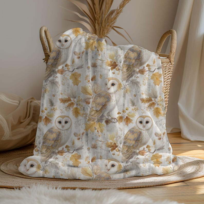 May include: A white and beige blanket featuring a repeating pattern of owls, branches, and autumn leaves. The blanket is folded in a woven basket. The owls have brown and white feathers. The leaves are shades of yellow and brown.