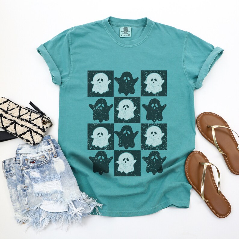 May include: Teal blue t-shirt with a checkered pattern of white and dark blue ghosts.