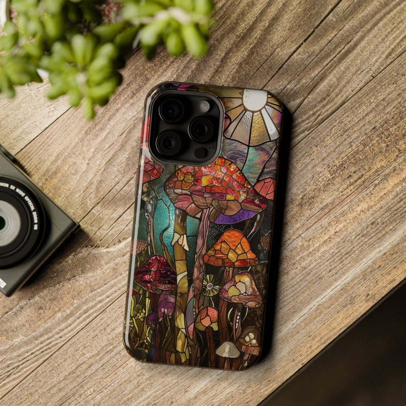 May include: A colorful phone case with a stained glass design featuring mushrooms and a sun. The case is made for an iPhone with a triple lens camera.