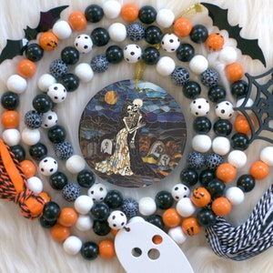 May include: A Halloween-themed beaded garland with orange, white, and black beads. The garland features a stained glass-style ornament depicting a skeleton couple embracing in a graveyard setting.