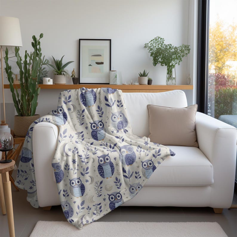 May include: A white throw blanket with a blue and white owl pattern. The owls are surrounded by stars, leaves, and crescent moons. The blanket is draped over a white couch.