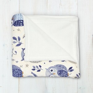 May include: A soft, white fleece blanket with a cream-colored background featuring a repeating pattern of blue owls, leaves, and stars. The blanket is folded in half, revealing the white fleece underside.