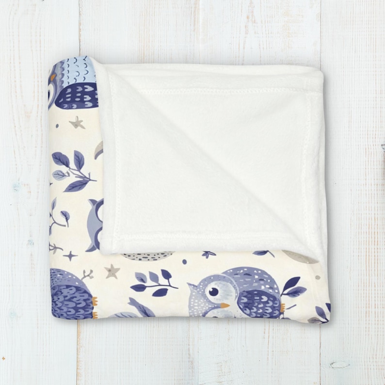 May include: A soft, white fleece blanket with a cream-colored background featuring a repeating pattern of blue owls, leaves, and stars. The blanket is folded in half, revealing the white fleece underside.
