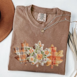 May include: A brown Comfort Colors t-shirt with three leaf appliqués. The leaves are patterned with plaid and floral designs in orange, yellow, and green. A silver necklace with a small pendant rests on the shirt. The shirt tag reads "S - P - CH".