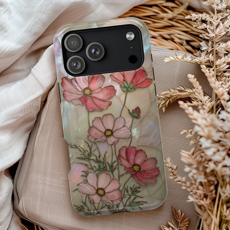 May include: A smartphone case featuring a floral design with pink cosmos flowers and green leaves. The case has a pearlescent background and a black camera module.
