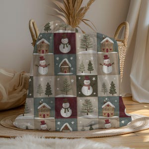 May include: A Christmas-themed throw blanket with a patchwork design. The blanket features squares with images of snowmen, houses, and Christmas trees in shades of red, green, and beige. The blanket is folded over a wicker basket.