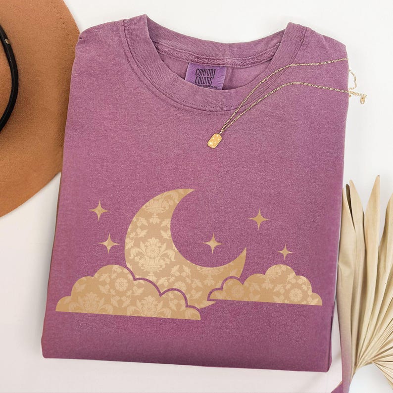 May include: A purple Comfort Colors t-shirt featuring a gold crescent moon and cloud design with stars. A gold necklace with a rectangular pendant rests on the shirt. A brown hat and dried palm leaves are in the background.