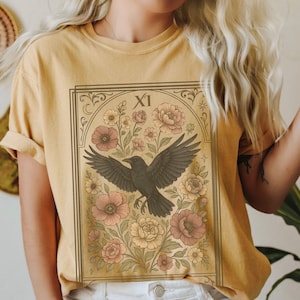 May include: A yellow t-shirt with a vintage tarot card design featuring a black crow flying over a floral arrangement. The card number "XI" is visible in the top right corner.