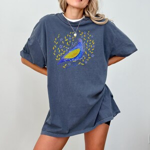 Puede incluir: Una camiseta azul oscuro con un dise&ntilde;o de p&aacute;jaro colorido. El p&aacute;jaro es azul y amarillo, rodeado por una corona de hojas amarillas y flores moradas. La camiseta tiene mangas cortas y un corte holgado.