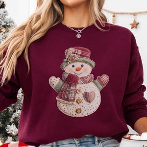 May include: A maroon sweatshirt featuring a snowman design. The snowman has a plaid hat, scarf, and mittens, with a heart-shaped patch and button details. The snowman's face has a carrot nose and a smiling expression. A festive winter-themed garment.