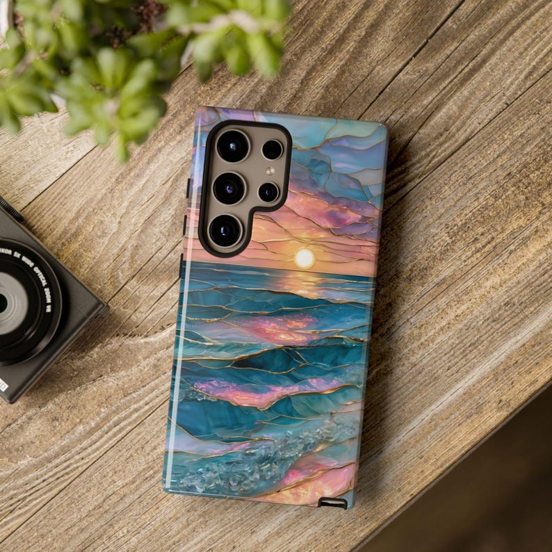 May include: A smartphone case featuring a sunset over the ocean design. The case has a colorful, artistic depiction of the sea with blue, pink, and gold accents. The phone case is on a wooden surface.
