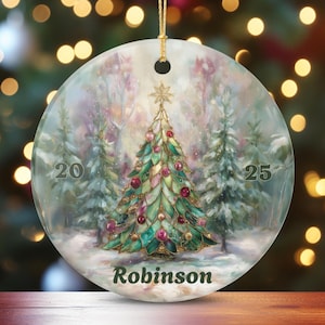 May include: A round, flat, glass-like Christmas ornament featuring a stained-glass style tree in shades of green and gold with red accents. The design includes a snowy forest background, the year 2025, and the name Robinson printed in dark text.