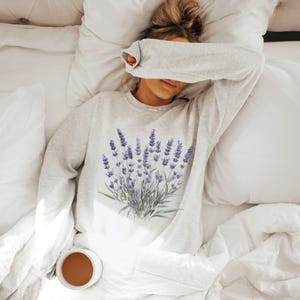 Op de afbeelding: Een grijs sweatshirt met een lavendelbloemenprint. Het sweatshirt wordt gedragen door iemand die op een wit bed ligt.