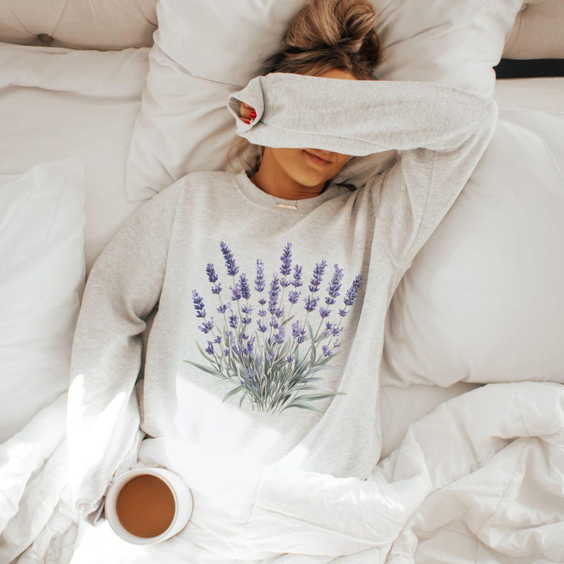 Op de afbeelding: Een grijs sweatshirt met een lavendelbloemenprint. Het sweatshirt wordt gedragen door iemand die op een wit bed ligt.