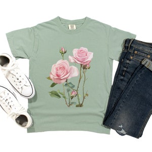 May include: A sage green t-shirt with a pink rose floral print. The shirt has a crew neck and short sleeves.