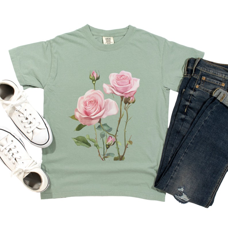 May include: A sage green t-shirt with a pink rose floral print. The shirt has a crew neck and short sleeves.
