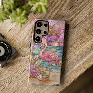 Puede incluir: Funda de tel&eacute;fono con un flamenco rosa en una playa con conchas y un atardecer. La funda tiene un efecto de vidriera en tonos rosa, morado y azul. Colocada sobre una superficie de madera.