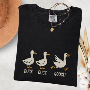 May include: Black t-shirt with a playful graphic of three ducks, with the text "DUCK DUCK GOOSE!". The ducks are white with orange beaks and feet. A necklace with a pendant is draped over the shirt.