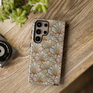 May include: A smartphone case with a pearlescent, fan-shaped pattern in shades of white, gold, and iridescent colors. The case has a camera module with multiple lenses. The phone case is on a wooden surface.