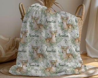 May include: A white blanket with a repeating pattern of deer wearing floral crowns, surrounded by green foliage and the name "Amelia" in black script. The blanket is folded in a wicker basket.